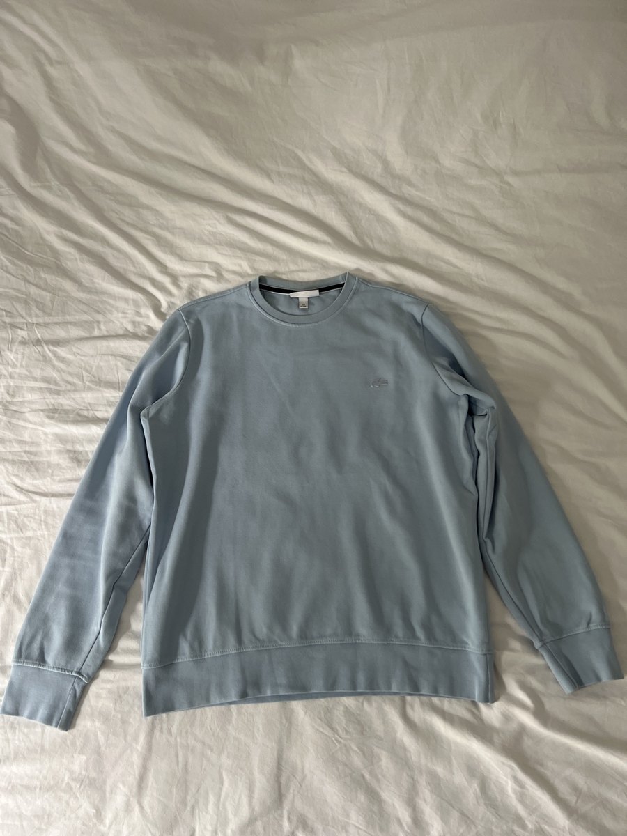 Un sweat bleu-gris photographié à plat sur un lit, en lumière d’intérieur.